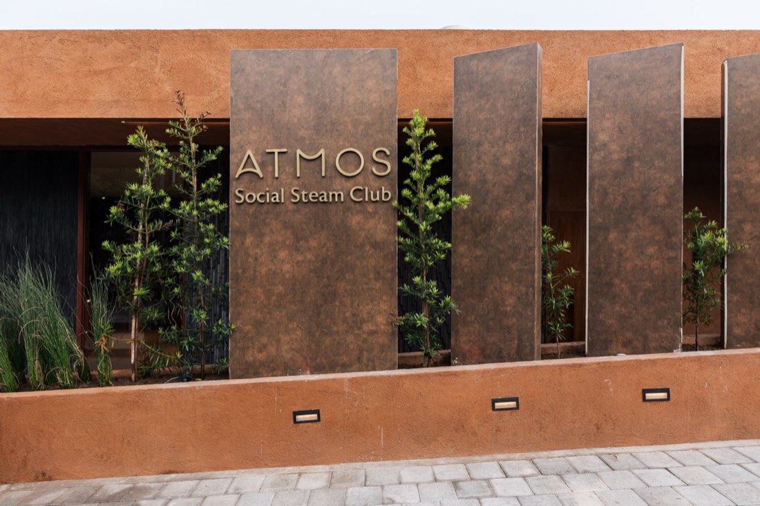 Facade of the ATMOS Social Steam Club showing a row of vertical patinated-metal panels and slender potted shrubs flanking the gold ATMOS Social Steam Club signage