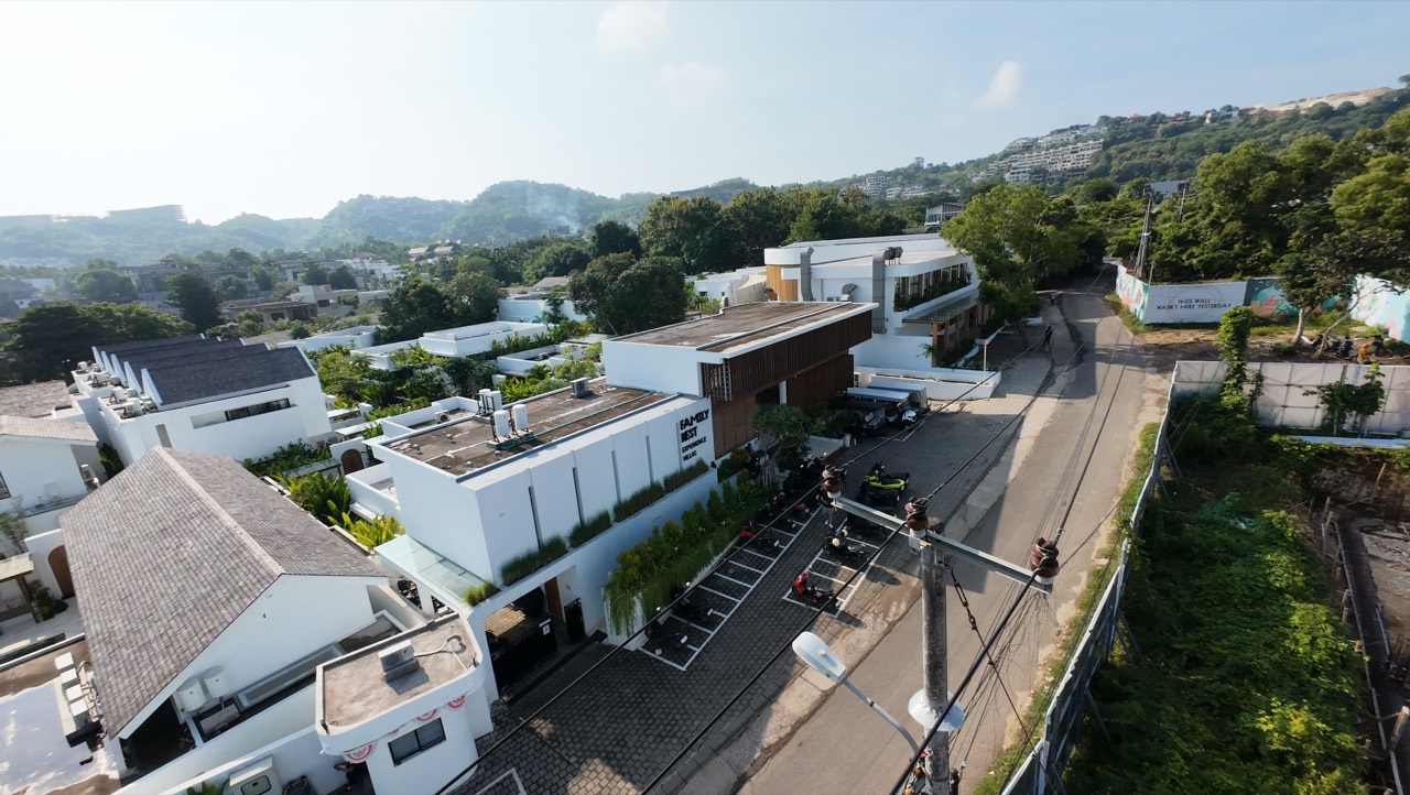 High aerial over the Family Nest resort and neighbouring white villas with rooftop terraces, the curved timber clubhouse, and a winding lane leading toward the green hilltops of Bali
