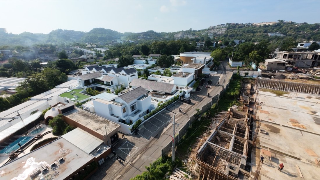 Wide aerial of the Family Nest resort district with rows of white villas and rooftop lawn terraces spread across a green Bali hillside under a dramatic blue and cloud-filled sky