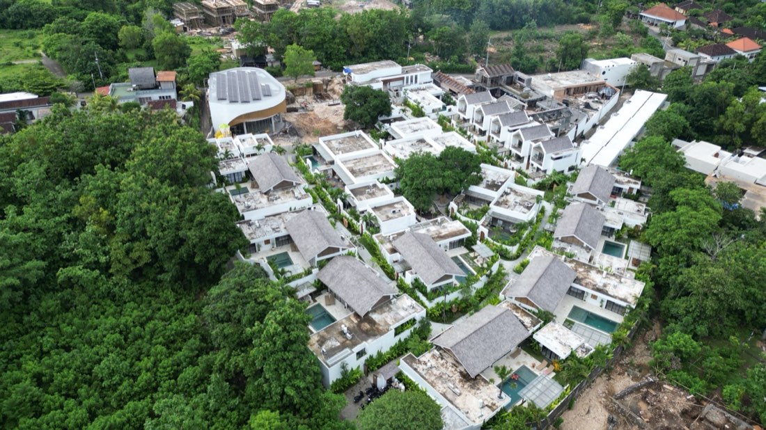 Aerial view of the Family Nest resort villas with grey thatched roofs, private pools and rooftop terraces on a green Bali hillside in Pecatu, Uluwatu