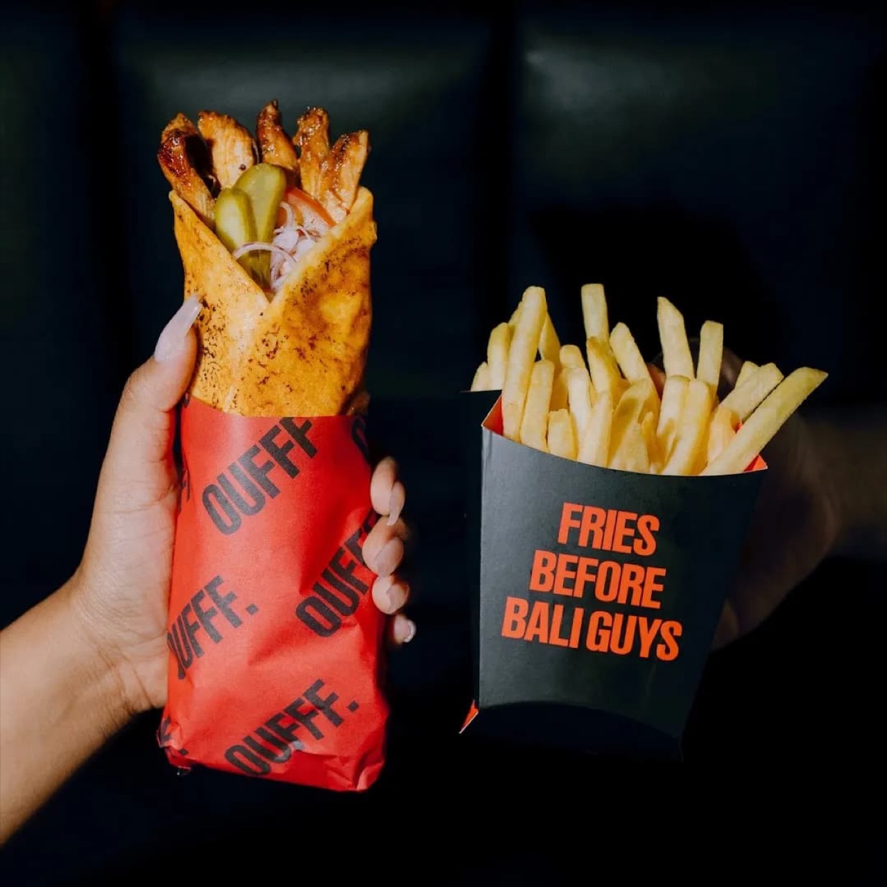 A hand holds a grilled chicken shawarma wrap in red OUFFF-printed paper beside a black fries box reading FRIES BEFORE BALI GUYS in orange, filled with golden french fries, against a dark background