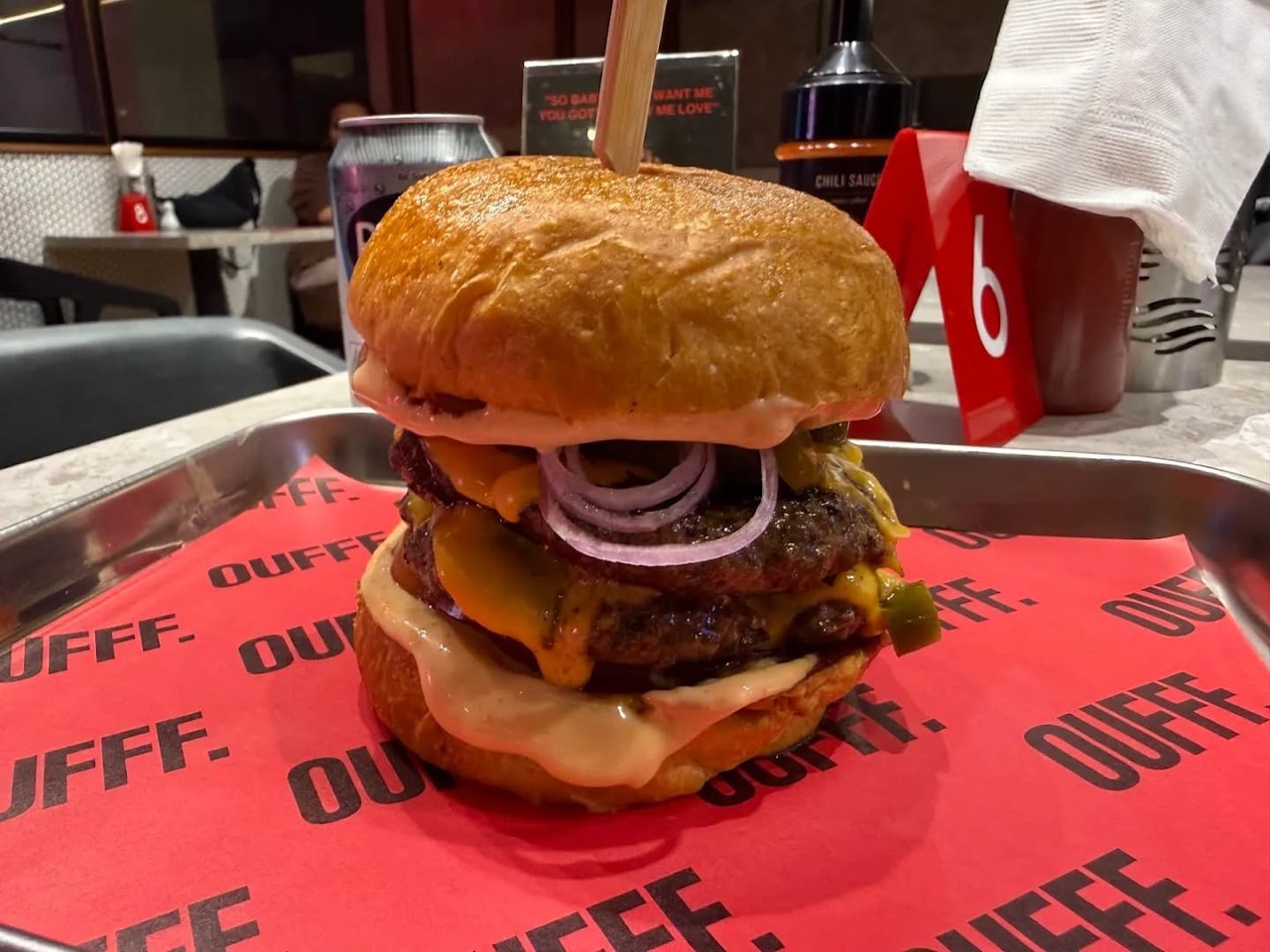 A tall double cheeseburger with a wooden pick on red OUFFF tray paper, dripping pink sauce with red onion rings and cheese, set on a metal tray with a soda can, chili-sauce bottle and napkin holder on the table behind