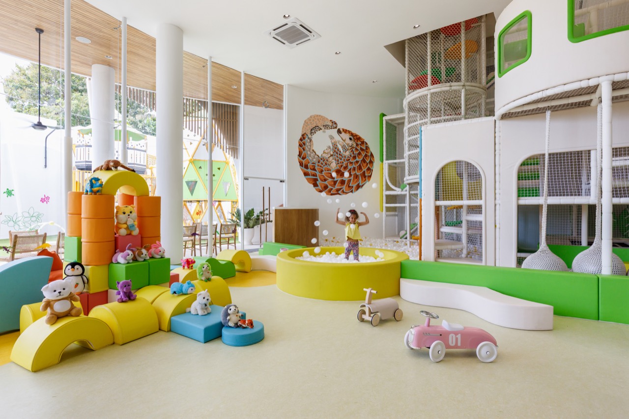 Bright play area inside the Pangolin Kids Club where a child plays in a round white ball pit beneath the pangolin mural, with soft foam blocks, plush toys, ride-on toy cars, and the two-storey play structure