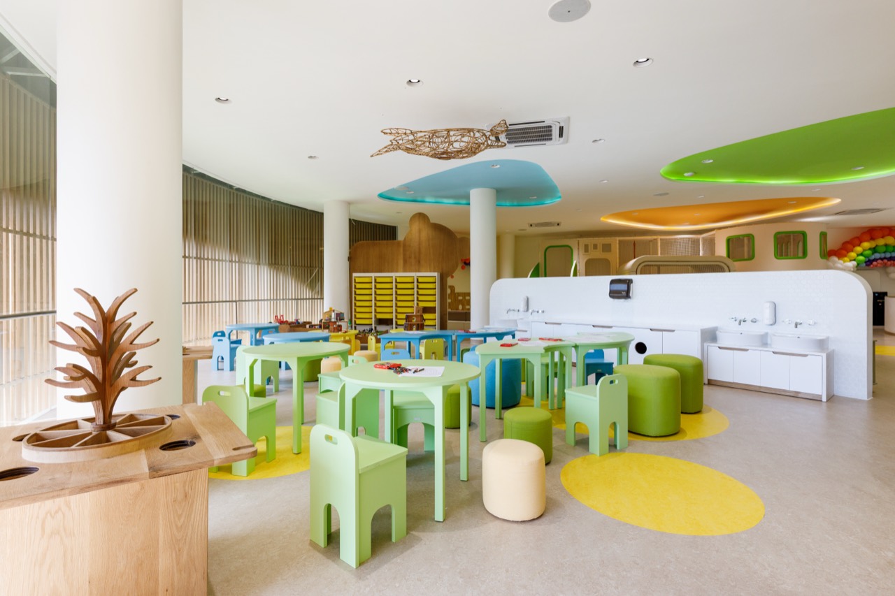 Craft and activity room at the Pangolin Kids Club with rounded green child-sized tables and chairs, a wooden play island, storage cubbies, sinks along the wall, and pastel ceiling discs