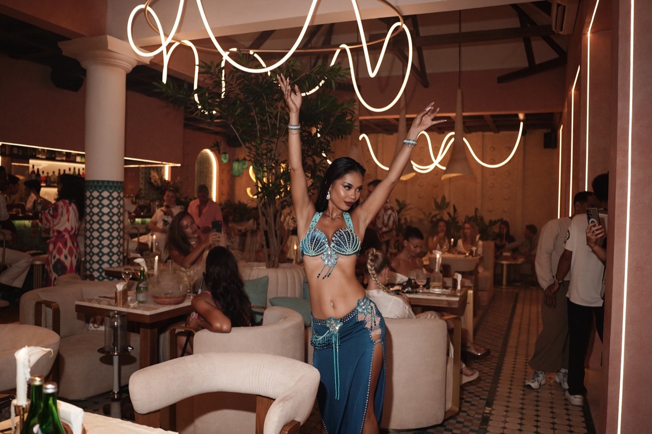 A belly dancer in a turquoise beaded bra and blue skirt performs with arms raised in a busy candlelit dining room with looping neon lights, plush beige seating and seated guests in the background