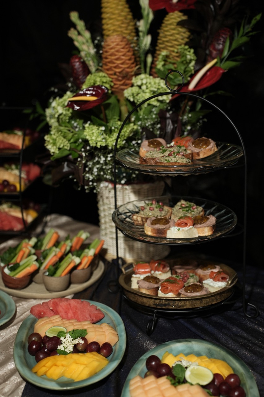 A three-tier glass stand of canapes topped with smoked salmon, cherry tomato and herbs, beside plates of sliced mango, melon and grapes and vegetable crudites, with a tall tropical flower arrangement behind on a dark cloth