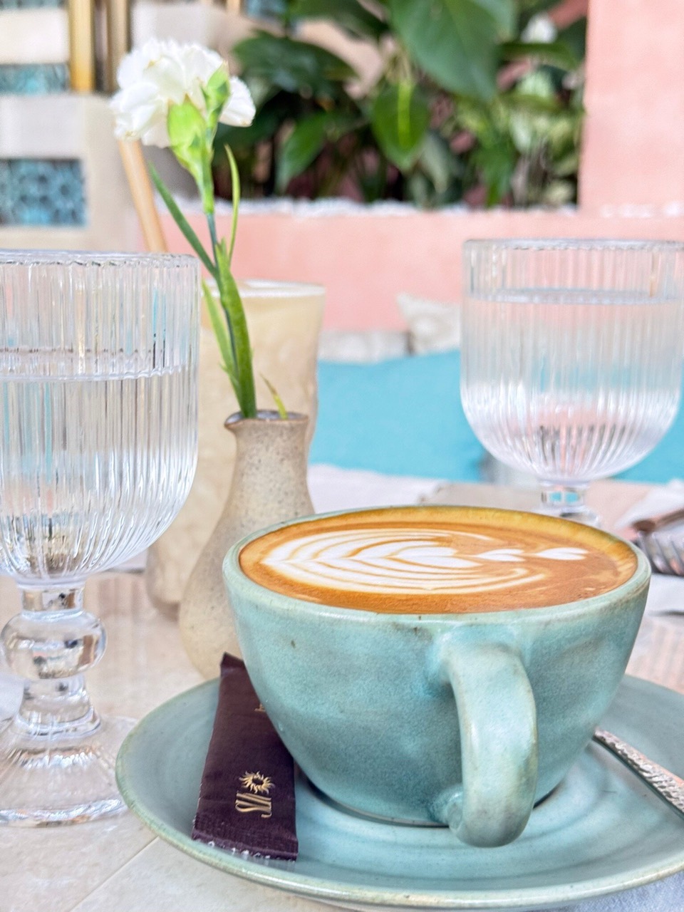 A latte with leaf rosetta art in a turquoise ceramic cup and saucer on a table with two ribbed glass tumblers of water and a small vase holding a white carnation, with green plants and a pink wall behind